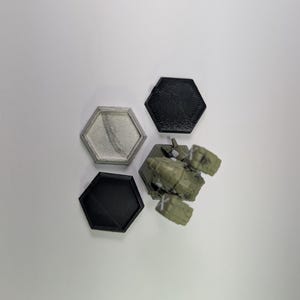 May include: An overhead shot shows four hexagonal objects. Two are black, one is silver with a textured interior, and the last is a green and brown object with a complex shape. The objects are arranged on a white surface.