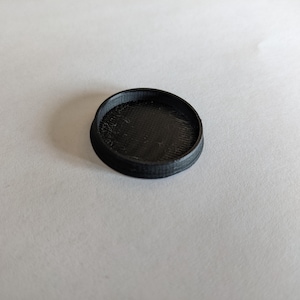 May include: A small, circular, black object with a textured inner surface and a raised rim. The item is made of a solid material and rests on a white surface. The design is simple and functional.