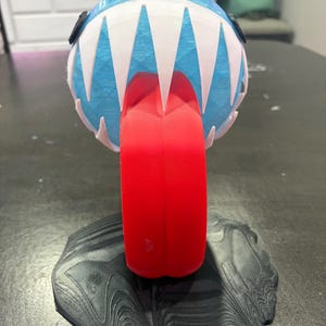 Könnte beinhalten: Eine 3D-gedruckte, kugelförmige Kreaturenfigur mit hellblauem Körper, spitzen weißen Zähnen und einer markanten roten Zunge, die nach unten ragt. Die Figur steht auf einem strukturierten, schwarzen, runden Sockel mit wellenförmigem Muster.