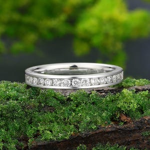 May include: A silver band ring featuring a continuous line of small, clear gemstones. The ring is presented on a bed of bright green moss, with a blurred green backdrop.