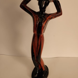 May include: A ceramic figurine of a person in a hat, with arms raised. The figure is primarily black with red and brown vertical stripes. The base is a black oval. The hat is brown.