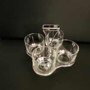 Peut inclure: Un service de présentation en verre transparent avec quatre petits verres et une poignée centrale. Les verres sont posés sur un socle en métal argenté. L'ensemble est conçu pour servir des condiments ou de petites portions.