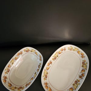 Puede incluir: Dos fuentes ovaladas con interior blanco y borde decorativo. El borde presenta un patrón repetido de peras amarillas, nueces marrones y hojas verdes. Las fuentes están sobre un fondo oscuro.