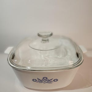 May include: White square casserole dish with a clear glass lid. The dish has a blue floral design on the front and white handles. A clear glass knob is on top of the lid.