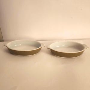 Puede incluir: Dos fuentes redondas para hornear. Cada fuente tiene un interior blanco y un exterior marrón claro. Las fuentes tienen asas pequeñas a cada lado. Las fuentes están vacías y sobre una superficie blanca.