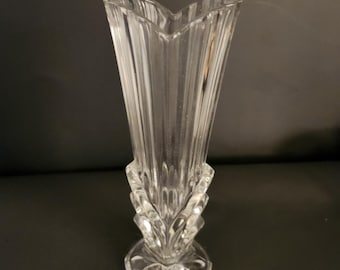 May include: A clear glass vase with a fluted, heart-shaped top and a geometric base. The vase has vertical ridges and a decorative, layered design at the base. The vase is designed to hold flowers.