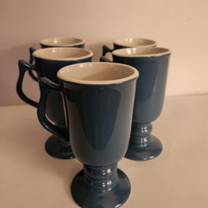 May include: Five blue ceramic mugs with white interiors and handles. The mugs have a pedestal base and a glossy finish. The mugs are arranged on a white surface.