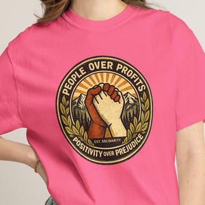 May include: A bright pink short-sleeve t-shirt featuring a circular graphic on the chest. The design shows two hands clasped together with the text "PEOPLE OVER PROFITS" and "POSITIVITY OVER PREJUDICE" surrounding a mountain landscape and laurel leaves.