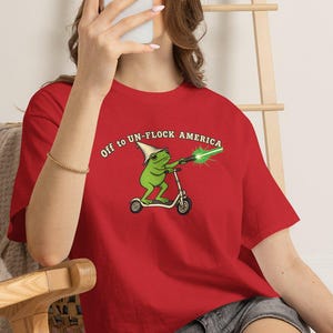 Może przedstawiać: Czerwony t-shirt z grafiką przedstawiającą zieloną żabę w czapce imprezowej, jadącą na hulajnodze i trzymającą świecący zielony miecz świetlny. Tekst nad żabą brzmi "Off to UN-FLOCK AMERICA" i jest napisany zakrzywioną, białą czcionką.
