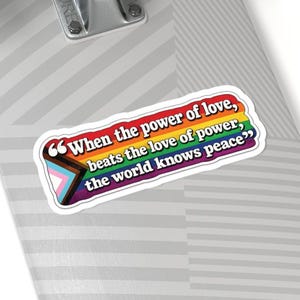 May include: A rectangular sticker with rounded corners featuring the text "When the power of love, beats the love of power, the world knows peace" in white letters. The background displays a rainbow pride flag with black, brown, blue, pink, and white stripes.