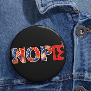 May include: A black button with the word "NOPE" in red, white, and blue, with a patriotic design. The button is round and is set against a blue denim jacket.