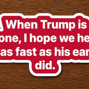 Op de afbeelding: Een rode sticker met witte tekst die luidt: "When Trump is gone, I hope we heal as fast as his ear did." De sticker heeft een witte rand en ligt op een bruin houten oppervlak.