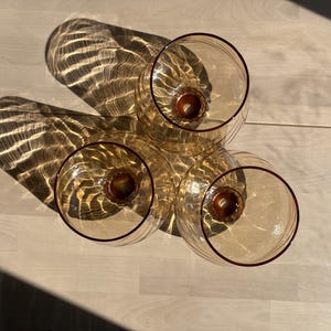 May include: Four amber-colored, stemmed glasses with a rounded bowl and a dark brown rim. The glasses are arranged on a light-colored wooden surface, casting elongated shadows. The sunlight creates a play of light and shadow.