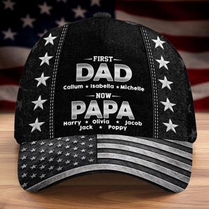 May include: Black baseball cap with an American flag design on the brim. The cap features the text "FIRST DAD NOW PAPA" with names of children. White stars are embroidered on the sides.