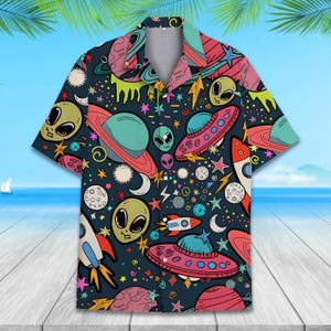 May include: Short-sleeved button-down shirt with an all-over space-themed print. The design features planets, rockets, UFOs, aliens, stars, and moons in vibrant colors on a dark blue background. The shirt has a classic collar and a relaxed fit.