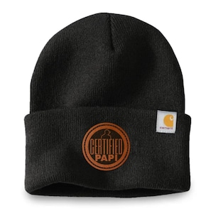 May include: Black Carhartt beanie with a brown leather patch that reads "CERTIFIED PAPI" in a circular design. The beanie has a folded cuff and a small Carhartt label. A warm winter hat.