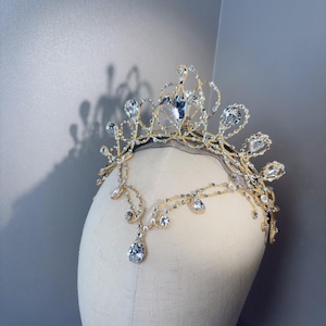 May include: A gold-toned metal tiara featuring a central elevated crown design and a delicate forehead chain. The piece is adorned with numerous clear, faceted crystals in various shapes, including teardrop and round cuts, set in a filigree pattern.