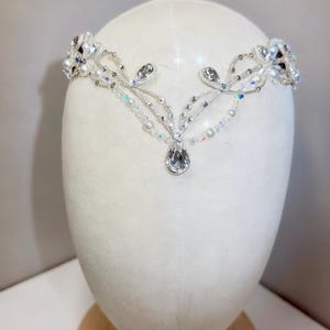 May include: A delicate silver-toned tiara with clear and iridescent crystals, pearls, and beads. The headpiece features a central teardrop-shaped crystal and floral-like accents on each side. The tiara is displayed on a white mannequin head.