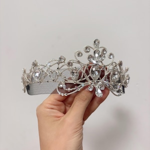 May include: A silver-toned tiara with clear, sparkling gemstones. The crown features an intricate design with a mesh base and is held by a hand against a white background. The gemstones are various shapes and sizes, adding to the tiara's elegance.