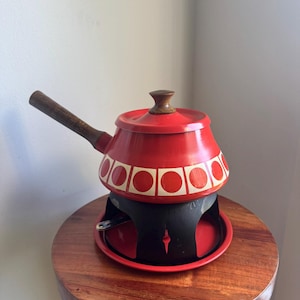 Puede incluir: Una olla para fondue de esmalte rojo con mango y pomo de tapa de madera. La olla presenta una banda blanca con círculos rojos alrededor de la base. Se asienta sobre un soporte de metal negro con una base circular roja para el calor.