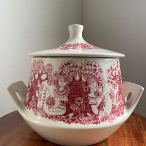 Könnte beinhalten: Weißer Keramik-Serviertopf mit Deckel und rotem Blumen- und Figuren-Design. Die Schüssel hat zwei Griffe und einen abgerundeten Boden. Der Deckel hat einen Knauf und ein passendes rotes Blumenmuster.