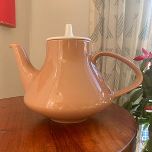 Może przedstawiać: Brzoskwiniowa ceramiczna czajniczek z białą pokrywką i uchwytem. Czajniczek ma zakrzywioną wylewkę i zaokrąglony korpus. Uchwyt to zakrzywiona pętla. Czajniczek stoi na drewnianym stole.