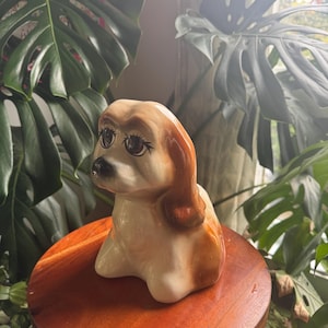 May include: A ceramic dog figurine featuring a glossy cream and brown finish. The dog has large, dark brown eyes with prominent painted eyelashes, a black nose, and long, smooth ears that drape down the sides of its head in a seated position.
