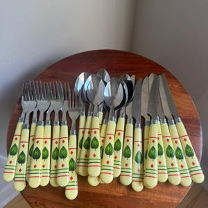May include: A collection of stainless steel flatware featuring cream-colored handles decorated with green leaf motifs, red diamond accents, and horizontal green lines. The set includes multiple forks, spoons, and knives with matching patterns.