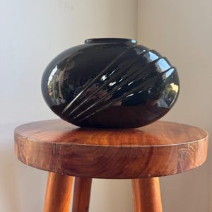 Peut inclure: Un vase en céramique noir brillant, de forme arrondie, avec un motif texturé en forme d'éventail sur un côté. Le vase est posé sur un petit tabouret rond en bois à trois pieds. Le vase reflète l'environnement.