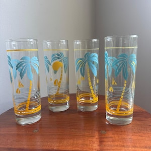 May include: A set of four clear glass tumblers featuring a tropical design with light blue palm trees, yellow suns, and small sailboats. Each glass has horizontal lines near the base and a thin yellow band around the top rim and the bottom section.