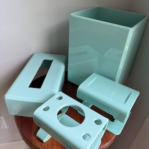 May include: A set of four light mint green bathroom accessories including a tall rectangular waste bin, a rectangular tissue box cover with a central opening, a soap dish, and a toothbrush holder with four circular openings for brushes.