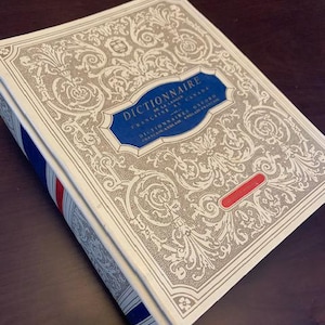 May include: A hardcover dictionary with a cream-colored cover featuring an ornate, embossed design. The title, "Dictionnaire," is displayed in a blue banner. The spine has red and blue stripes. The book is angled on a dark wooden surface.