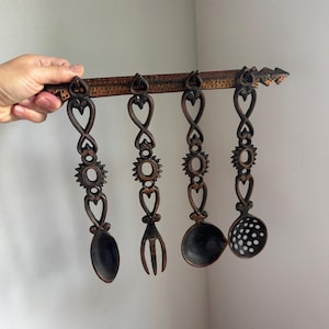 Peut inclure: Un support mural pour ustensiles de cuisine composé d'une barre en métal brun foncé texturé avec quatre outils suspendus. L'ensemble comprend une cuillère pleine, une fourchette à trois dents, une petite louche et une écumoire perforée.
