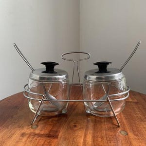 May include: A vintage glass condiment set with two clear jars, each with a silver lid and a black knob. The jars sit in a silver metal holder with a handle. Each jar has a small spoon inside.