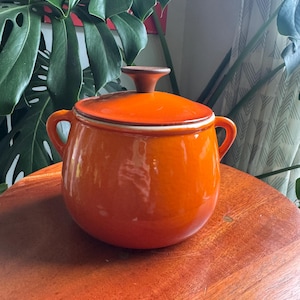 Peut inclure: Un pot en céramique orange vif avec un couvercle assorti et deux poignées latérales. Le pot présente une finition lisse et brillante ainsi qu'un bouton circulaire marron sur le dessus du couvercle. Le design est arrondi et bombé.