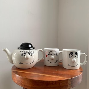 Peut inclure: Un ensemble de thé fantaisiste comprenant une théière blanche avec un chapeau noir et deux tasses assorties. Chaque pièce présente un visage souriant avec des détails de nez et d'yeux en relief. L'ensemble est présenté sur un tabouret en bois.
