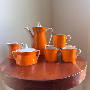 May include: A vintage tea set featuring a tall orange teapot with a white lid, a matching orange creamer, a lidded orange sugar bowl with two handles, and four orange tea cups. Each piece has a smooth, glossy finish and white interior accents.