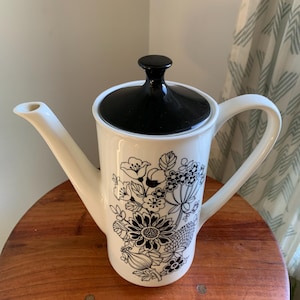 Op de afbeelding: Een witte keramische theepot met een zwarte deksel en handvat. De theepot is versierd met een zwart bloemenpatroon. De theepot staat op een houten oppervlak.