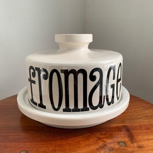 May include: A white ceramic cheese dish with a lid and a base. The word "fromage" is printed in large, black letters around the side of the dish. The dish is sitting on a wooden surface.