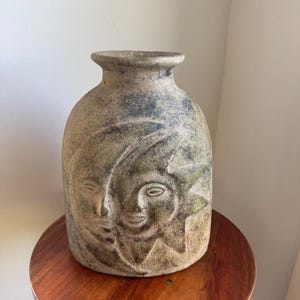 May include: A decorative vase featuring a sun and moon face design. The vase has a muted green and tan colour, with a rounded body and a narrow neck. The sun and moon faces are carved into the surface.
