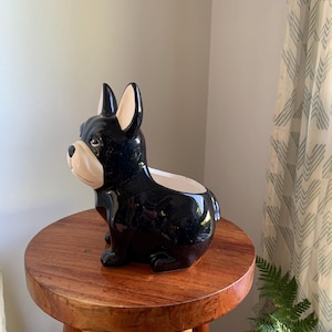 Könnte beinhalten: Ein schwarzer Keramik-Bulldoggen-Pflanztopf mit glänzendem Finish. Der Pflanztopf steht auf einem kleinen, runden Holzschemel. Der Hunde-Pflanztopf hat ein weißes Gesicht und Ohren und eine kleine Öffnung oben für Pflanzen.