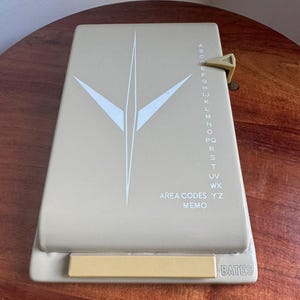 May include: A beige Bates telephone index with a rectangular base and a sliding selector tab. The top cover features a white geometric star-like graphic, a vertical alphabet list from A to Z, and the printed text AREA CODES MEMO and BATES.