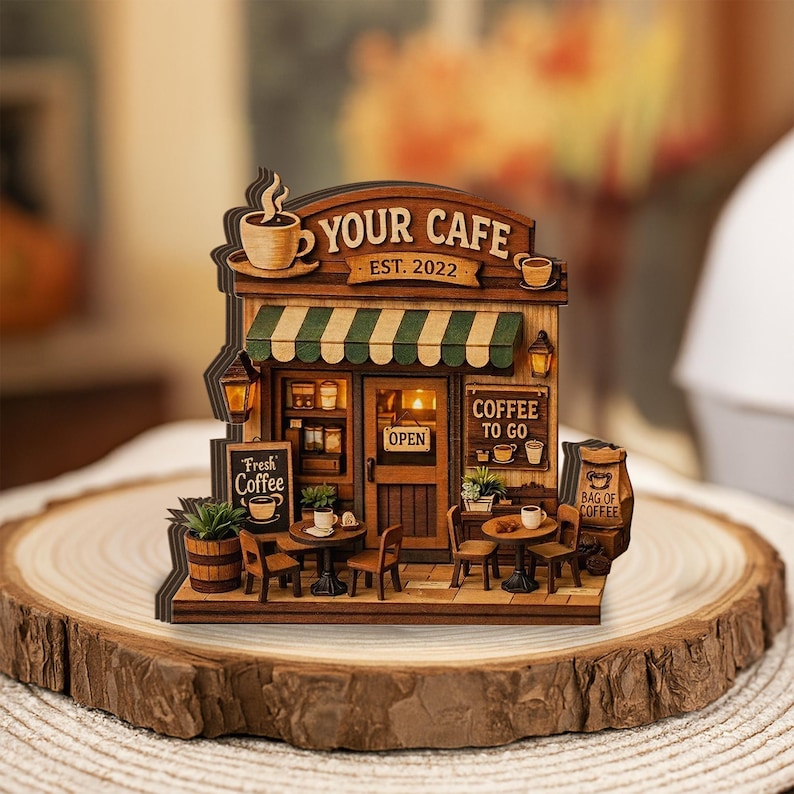 May include: A wooden model of a cafe with the text "YOUR CAFE EST. 2022". The miniature cafe has a green and white striped awning, small tables, chairs, and signs that read "Fresh Coffee" and "Coffee To Go".
