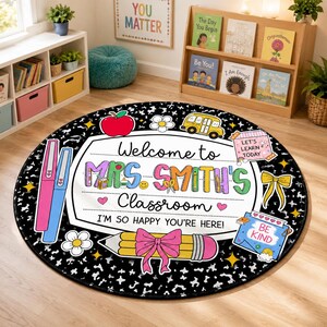 May include: A round classroom rug with a black and white speckled background. The center features a white sign with the text "Welcome to MRS. SMITH'S Classroom I'M SO HAPPY YOU'RE HERE!" decorated with a red apple, yellow bus, pencil, and bow.