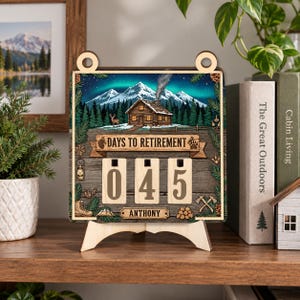 May include: A square wooden countdown sign featuring a mountain cabin scene with the text DAYS TO RETIREMENT and ANTHONY. The sign has three rotating number blocks currently displaying 045, decorated with pine trees, a deer, and axes.