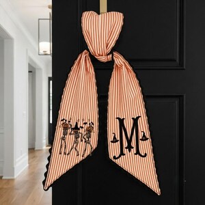 May include: A long, orange and white striped fabric door hanger with a black scalloped edge. The left tail features three dancing skeletons with pumpkin heads, while the right tail displays a large black letter M flanked by two witch hats.