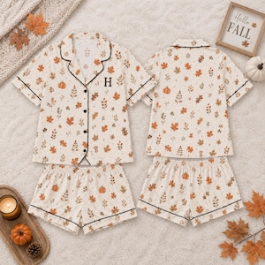 May include: A two-piece pajama set featuring a short-sleeved button-down shirt and matching shorts. The fabric is cream-colored with an all-over print of orange pumpkins and brown autumn leaves. The shirt features a black letter H and black piping.