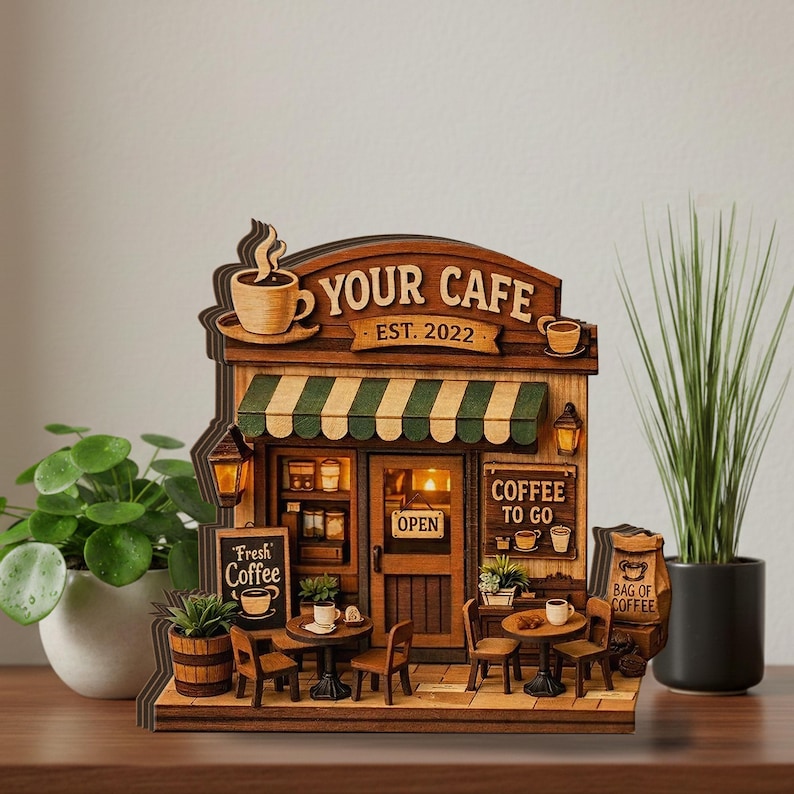 May include: A wooden model of a cafe, featuring the text "YOUR CAFE EST. 2022". The model has a green and white striped awning, small tables, chairs, and signs that read "Fresh Coffee" and "Coffee To Go".