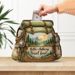 May include: A decorative backpack-shaped money bank with a scenic landscape painting. The bank is green and brown with the text "Bella + Anthony travel fund". A hand is inserting a $100 bill into the slot.