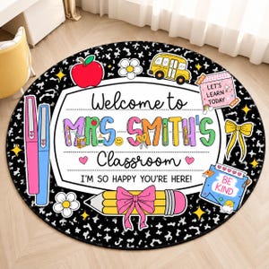 May include: A round classroom rug with a black and white speckled background. The center features a white sign reading Welcome to MRS. SMITH'S Classroom I'M SO HAPPY YOU'RE HERE! Decorated with a red apple, yellow bus, pencil, and notes.